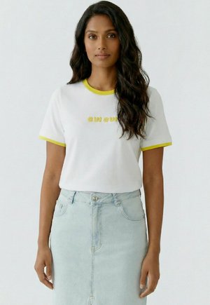 Frau mit langen, welligen Haaren, die ein weißes T-Shirt mit gelber Einfassung und dem Schriftzug "oui oui" trägt, kombiniert mit einem hellblauen Jeansrock.