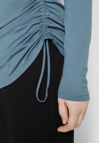 Langärmliges Oberteil aus weichem blauen Stoff mit gerafften Details an der Seite und einem Bindeband am Saum, kombiniert mit eng anliegenden schwarzen Hosen.