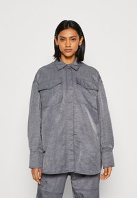 Grå oversized skjorte med krage, to brystlommer og lange ermer med knapper; laget av teksturert stoff.