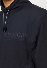 Sudadera azul marino con cuello alto, que incluye un cordón con regulador de plástico y el logo "HILFIGER" en relieve en el pecho. Textura suave.