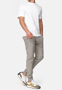 Homme portant un t-shirt blanc uni à manches courtes, un pantalon de jogging marron clair et des baskets blanches avec des accents jaunes et gris, se tenant avec une main dans sa poche.