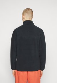 Giacca di pile nera con colletto alto, presenta una texture liscia e un taglio dritto, vista dal retro. Indossata con pantaloni arancioni larghi.