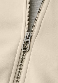 Fermeture éclair en métal avec gravure "STREET ONE" sur tissu beige, partiellement ouverte pour révéler un tissu gris clair en dessous.