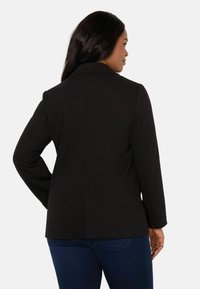 Black blazer with long sleeves, smooth texture, notch lapel, and back vent detailing. Paired with a dark denim outfit.