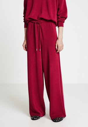 Rote Hose mit weitem Bein aus weichem Stoff, mit elastischem Bund und Kordelzug. Kombiniert mit gemusterten schwarzen Schuhen mit silbernen Akzenten.