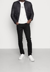 Svart vadderad jacka med ståkrage, vit T-shirt, svarta jeans och vita sneakers. Har logotyp på skjortan.