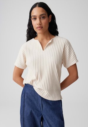 Donna con capelli lunghi e scuri che indossa una maglia crema a maniche corte a coste e pantaloni blu a vita alta, in piedi con le mani dietro la schiena.