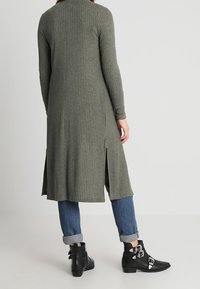 Abito a maniche lunghe a coste in verde oliva, con spacchi laterali e vestibilità comoda, abbinato a stivali neri alla caviglia. Jeans arrotolati alle caviglie.