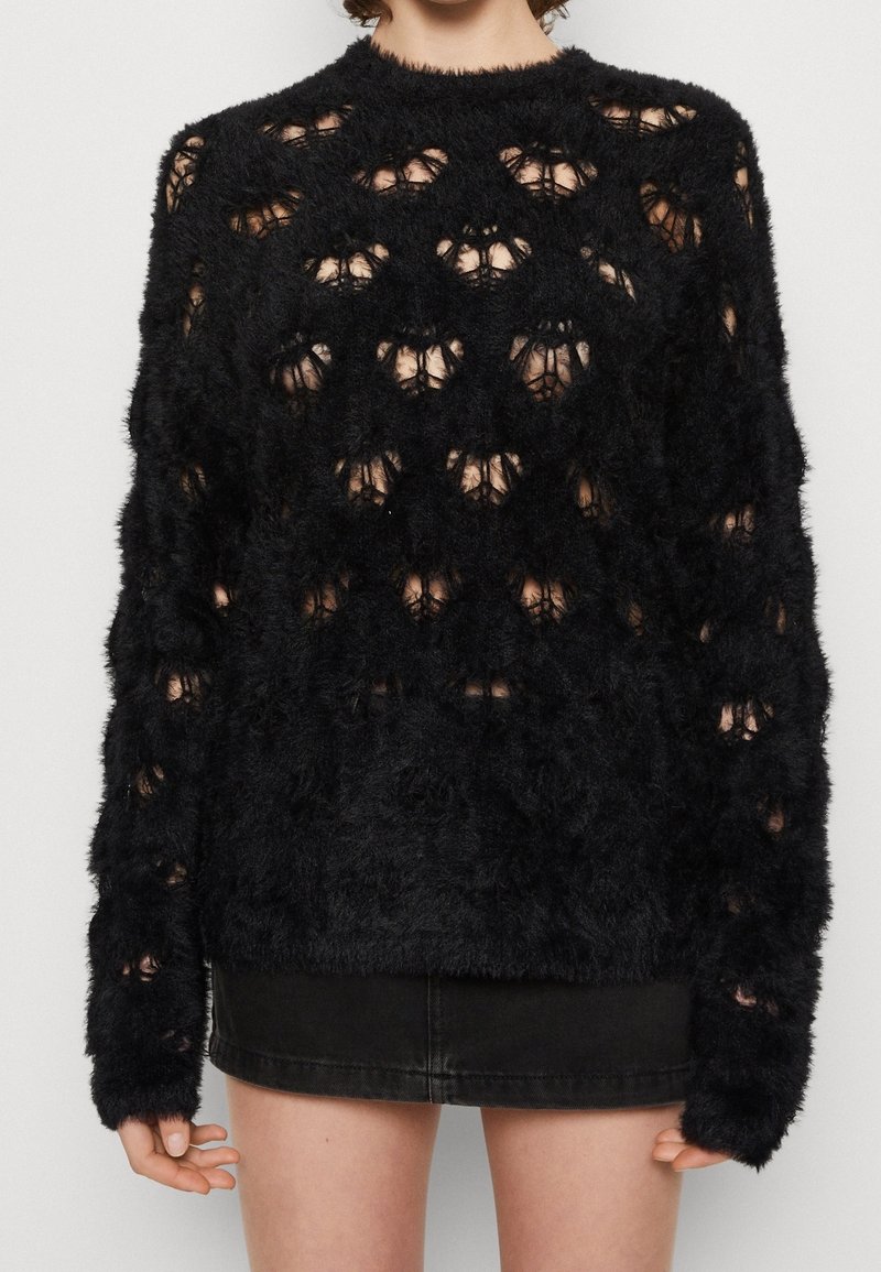 Personne portant un pull noir duveteux avec des trous irréguliers et une mini-jupe noire, debout devant un fond clair uni.
