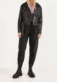 Vrouw in een zwarte leren motorjack, hoge zwarte joggerjeans, robuste zwarte laarzen en een lavendel ribbed top.