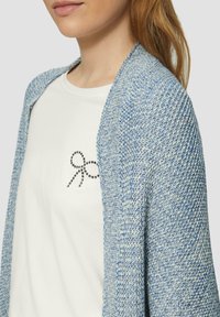 Cardigan en tricot bleu avec un motif texturé et une ouverture à l'avant, superposé sur un t-shirt blanc avec un détail de nœud clouté noir.
