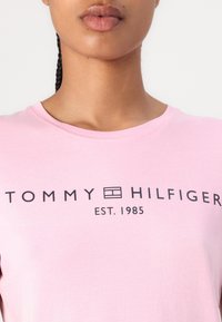 Gros plan d'une personne portant une chemise rose clair avec le logo noir « Tommy Hilfiger Est. 1985 » sur la poitrine.