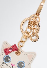 Portachiavi a forma di gatto realizzato in pelle crema con occhi blu e un fiocco rosa, dotato di accessori e dettagli in metallo dorato.