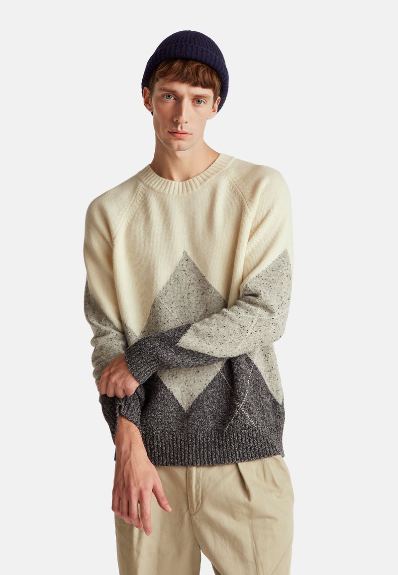 Kremowy i szary sweter dzianinowy z diamentowym wzorem. Prążkowany dekolt i mankiety. Modelka nosi ciemnoniebieską czapkę i beżowe spodnie.