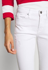 Jeans blancos con una textura suave, que presentan una pequeña área desgastada en el muslo, complementados por un cierre de botón clásico y cinco bolsillos.