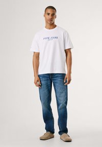 Camiseta blanca de algodón con letras azules "PEPE JEANS LONDON", combinada con jeans de mezclilla azul y zapatos tipo slip-on beige.