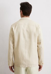 Helles beige Langarmhemd mit Kragen, das über eine glatte Textur, Knopfmanschetten und eine einzelne Naht entlang der Mitte des Rückens verfügt.