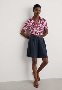 Florals on a short-sleeve, red, collared shirt; navy pleated shorts; brown braided belt; brown strappy sandals; neutral background.