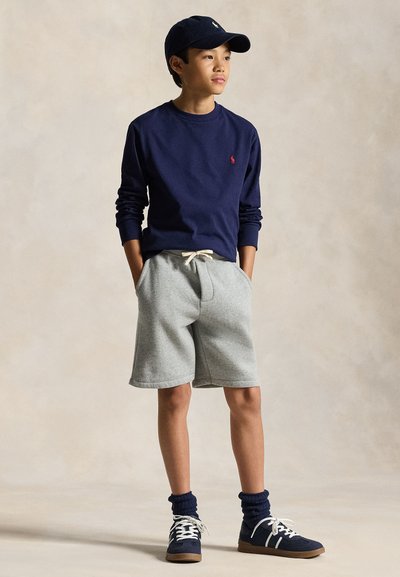 Teen boy wearing navy cap, navy sweatshirt, gray drawstring shorts, navy socks, and sneakers, standing with hands in pockets.