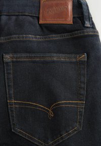 Mørke denimjeans har et lædermærke på taljen og kontrastfuld gul syning på baglommen med et buet sømdesign.