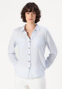 Button-up shirt in light blue striped fabric, featuring two breast pockets, long sleeves, and a collar. Textured and lightweight design.