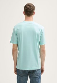 Lyseblå kortærmet t-shirt med rund hals og en glat tekstur, med et mærke på venstre ærme. Båret med blå jeans.