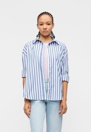 Ung kvinde med flettet hår iført en blå og hvid skjorte med lodrette striber over en lyse lilla top og lyseblå jeans.