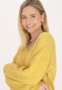 Geel gebreide trui met V-hals, met een zachte textuur en een relaxte pasvorm. Accessoires zijn ronde, lichtgekleurde oorbellen.