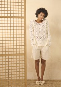 Witte haaktruien met ronde patronen, gecombineerd met beige shorts en lichte schoenen, tegen een neutrale achtergrond.