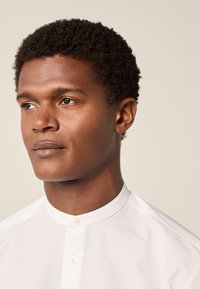 Bijela košulja s ovratnikom i visokim vratom; glatka tekstura; dva gumba na vratu; model s kratkom, kovrčavom tamnom kosom i neutralnom pozadinom.