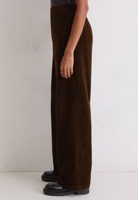 Bruine corduroy wijde beenbroek met een hoge tailleband, voorzien van een verticale ribbelstructuur en zwarte platform schoenen. Zijkantweergave tegen een neutrale achtergrond.