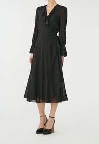 Schwarzes Kleid mit gerafftem Ausschnitt, langen Ärmeln und ausgestelltem Saum. Verziert mit einem Polka-Dot-Muster und kombiniert mit schwarzen High Heels.