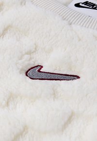 Nike fleeceová tkanina v off-white s plyšovou textúrou, s výšivkou šedého a vínového loga. Hladký golier pridáva detail.