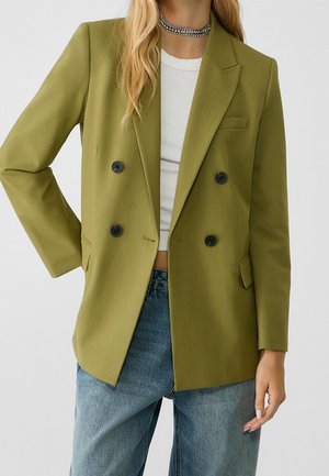 Vrouw draagt een olijfgroene double-breasted blazer, wit cropped topje, blauwe jeans en een sprankelende choker ketting tegen een effen achtergrond.