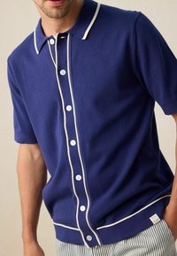 Navyblauw polo shirt met korte mouwen en witte bies, vijfknopenfront en ribgebreide onderkant. Gemaakt van zacht breiwerk met een gladde textuur.