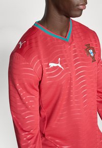 Κόκκινη ποδοσφαιρική φανέλα με λαιμόκοψη V, τυρκουάζ γιακά και οριζόντια ριγέ υφή. Διαθέτει λευκό λογότυπο Puma και το έμβλημα της Πορτογαλίας στο στήθος.