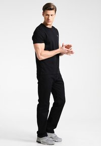 Man wearing black t-shirt and jeans, clasping hands, standing on a white background, facing slightly left.
