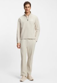 Beiger Pullover mit halbem Reißverschluss am Kragen und Logodetail; kombiniert mit passenden Hosen mit abgesteppten Nähten und geradem Bein.