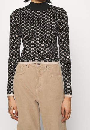 Nainen, jolla on musta istuva poolopaita, jossa on beige geometrinen kuvio, ja beige korkeavyötäröiset korduroihousut, seisoo valkoista taustaa vasten.