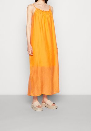 Robe maxi orange avec un col à volants et des fines bretelles. Tissu doux et léger créant une silhouette fluide. Associée à des sandales compensées beiges.