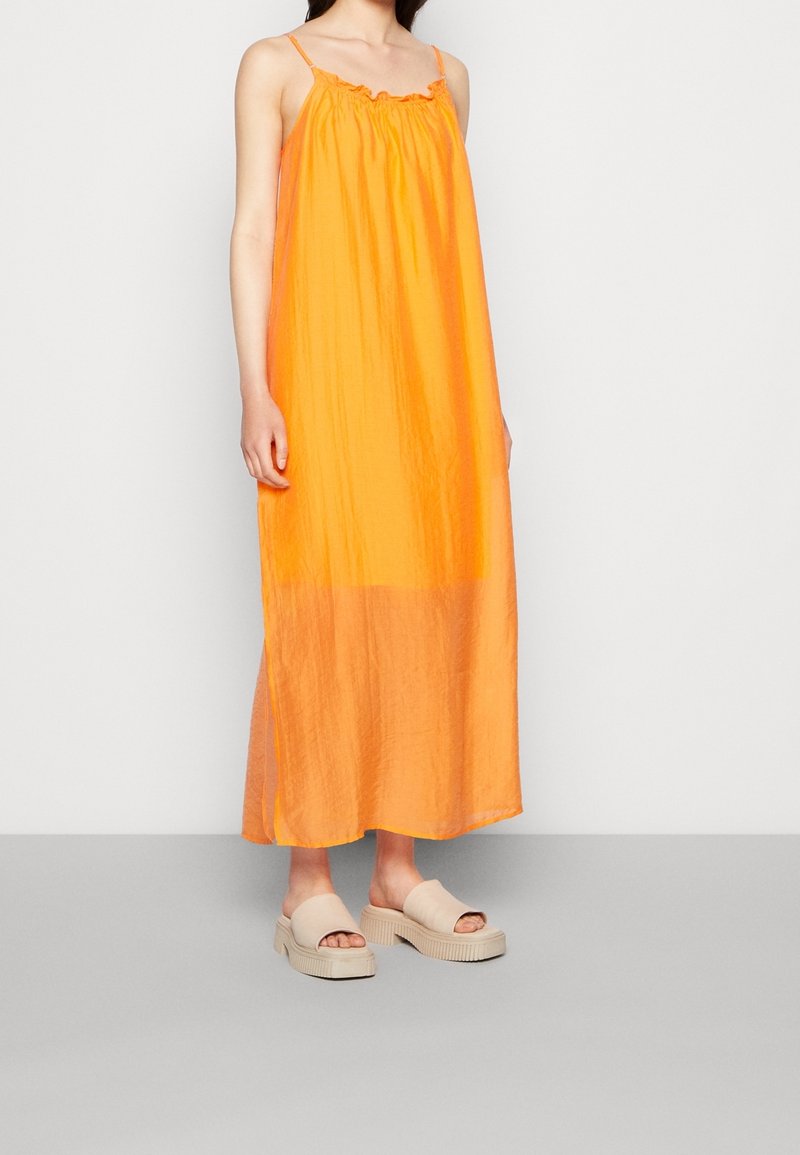 Robe maxi orange avec un col à volants et des fines bretelles. Tissu doux et léger créant une silhouette fluide. Associée à des sandales compensées beiges.