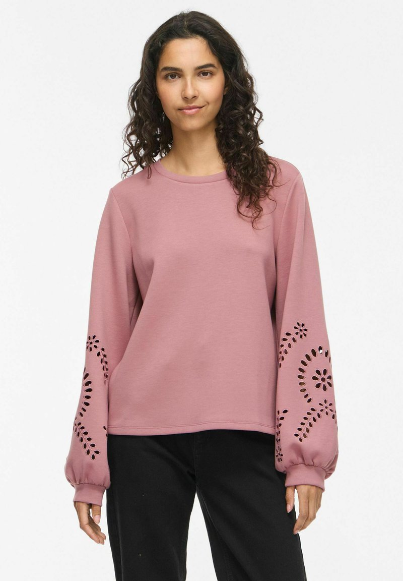 Femme aux cheveux bouclés portant un haut rose à manches longues avec des motifs floraux ajourés sur les manches et un pantalon noir.