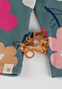 Baumwollkleidung mit einem türkisfarbenen Hintergrund, der große rosa und beige Blumenmuster zeigt. Befestigt ist ein braunes, pferdeförmiges Verzierungsstück.