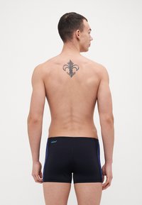 Sorte svømmebukser med blå sidestrehker, tætsiddende design og elastisk talje, med en synlig tatovering på den øvre del af ryggen.