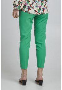 Green tapered pants with a smooth texture, ankle length, and a fitted design, paired with black high-heeled sandals.