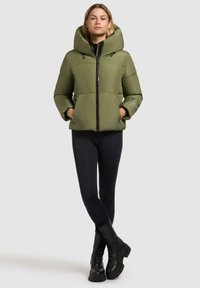 Olivengrøn pufferjakke med en stor hætte, lynlås lukning og to frontlommer. Sammen med sort leggings og combat støvler.