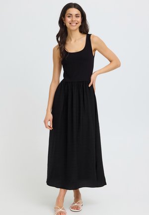 Femme souriante aux cheveux foncés portant une robe maxi noire sans manches et des talons blancs à brides, debout avec une main sur la hanche.