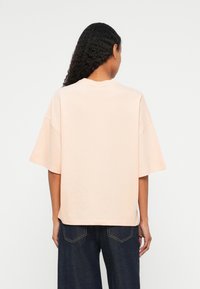 T-shirt oversized em rosa claro, com mangas curtas, decote redondo e uma textura suave. As costas são simples, sem detalhes de design visíveis.