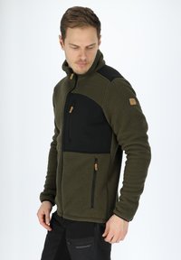 Giacca in pile di colore verde scuro con pannelli neri, tasca sul petto con zip e dettagli in metallo marrone. Colletto alto e tessuto texturizzato.