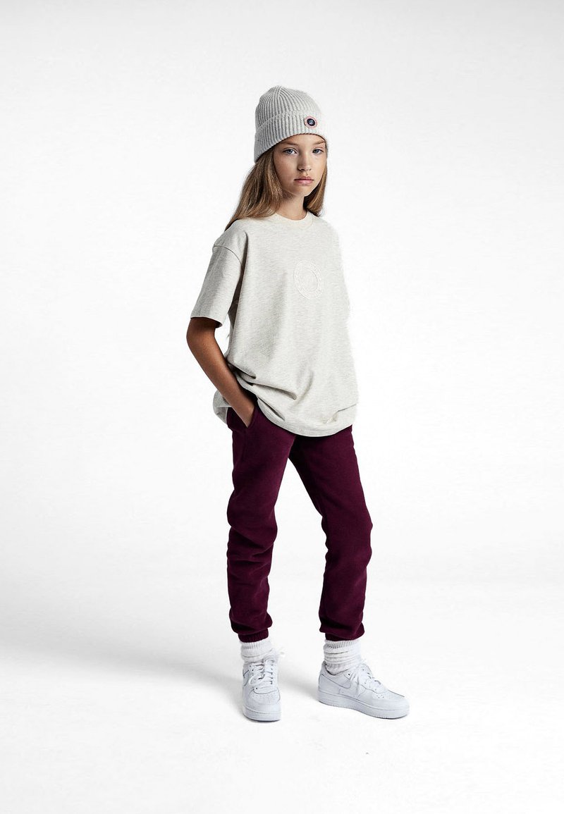 T-shirt oversize gris clair avec un logo rond, pantalon de survêtement bordeaux, baskets blanches et un bonnet côtelé gris clair. Tenue décontractée.